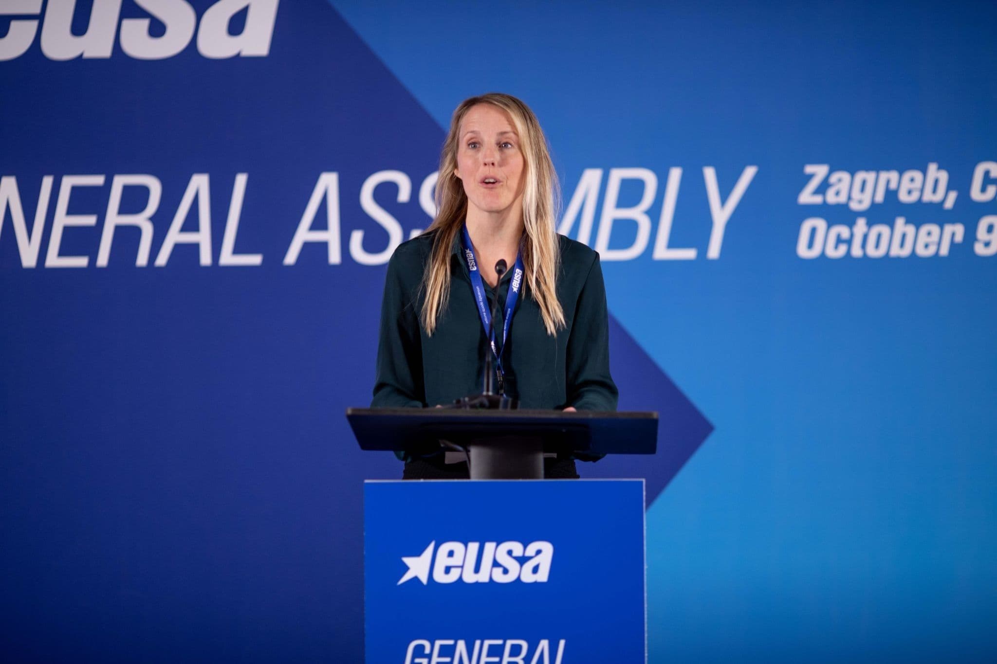 En kvinne holder tale bak en talerstol med EUSA-logo under en generalforsamling.