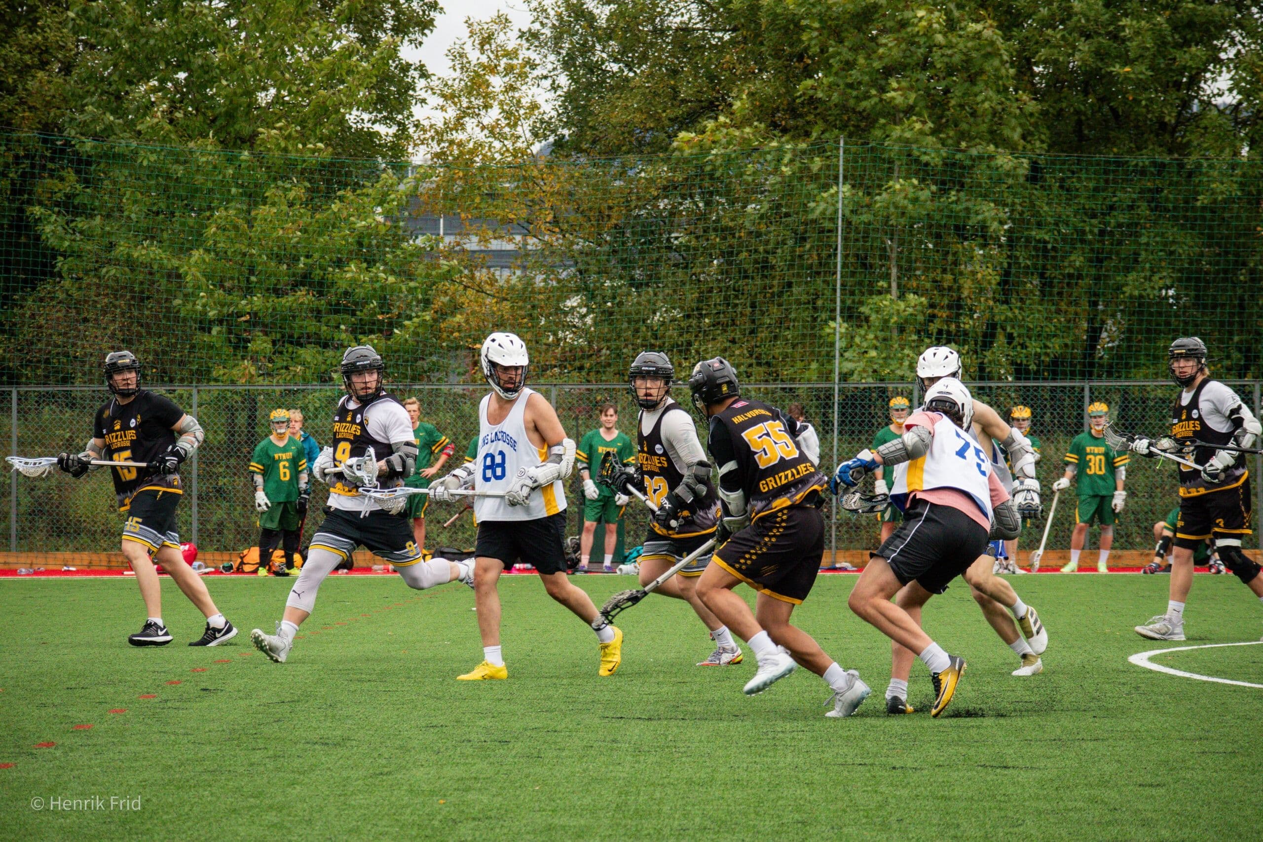 Lacrossekamp på kunstgress der spillere med køller og hjelmer kjemper om ballen.