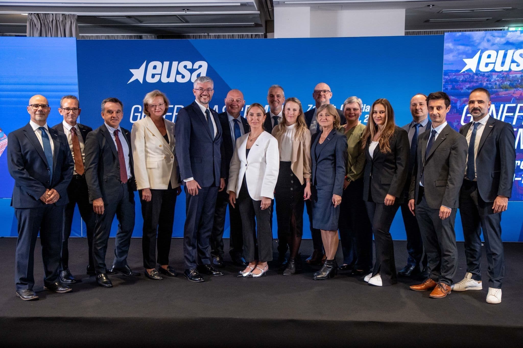 En gruppe mennesker i formelle klær poserer foran en EUSA-vegg under en generalforsamling.