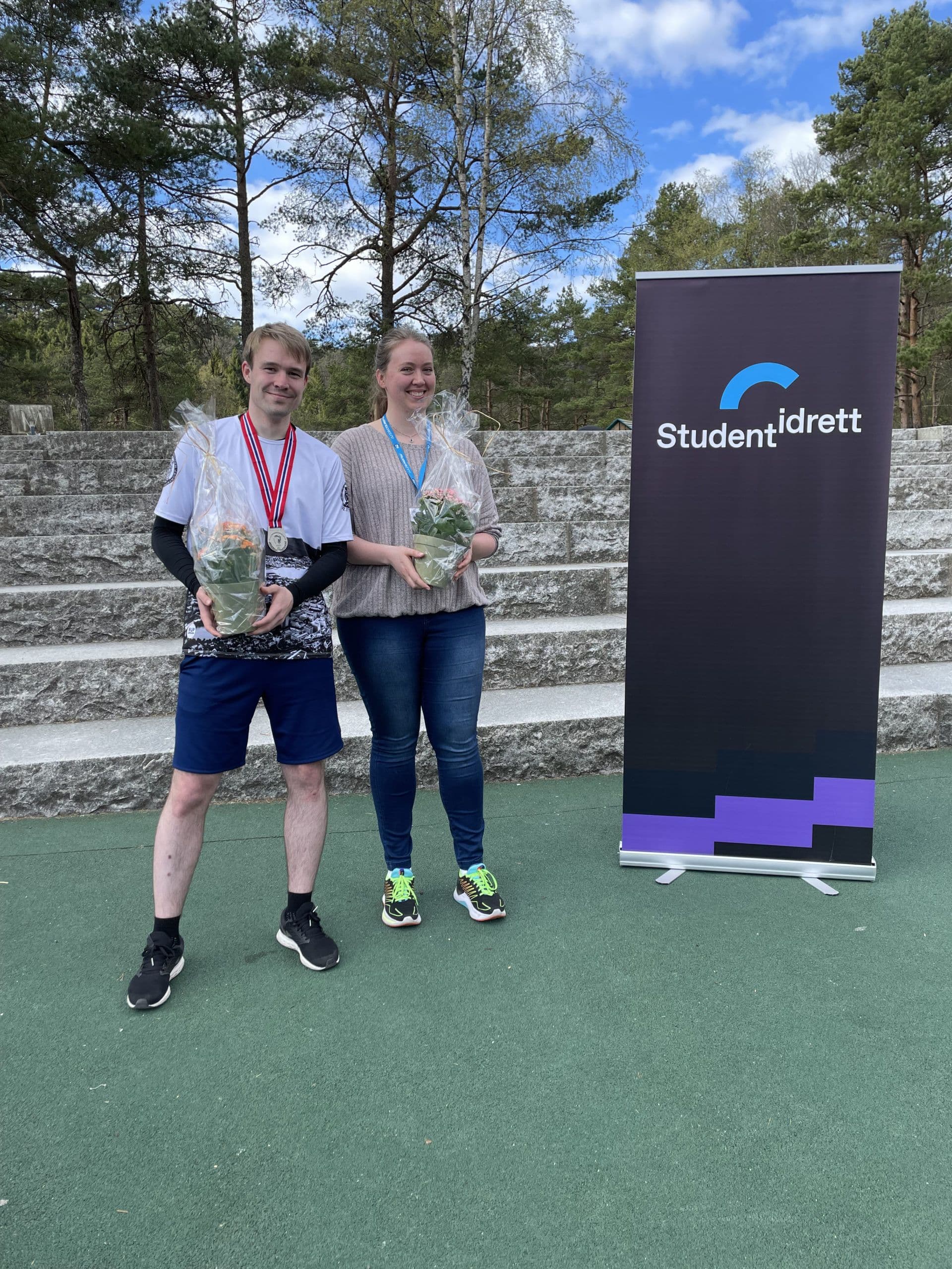 En mann og en kvinne med medaljer og blomster står ved siden av en Studentidrett-rollup utendørs.