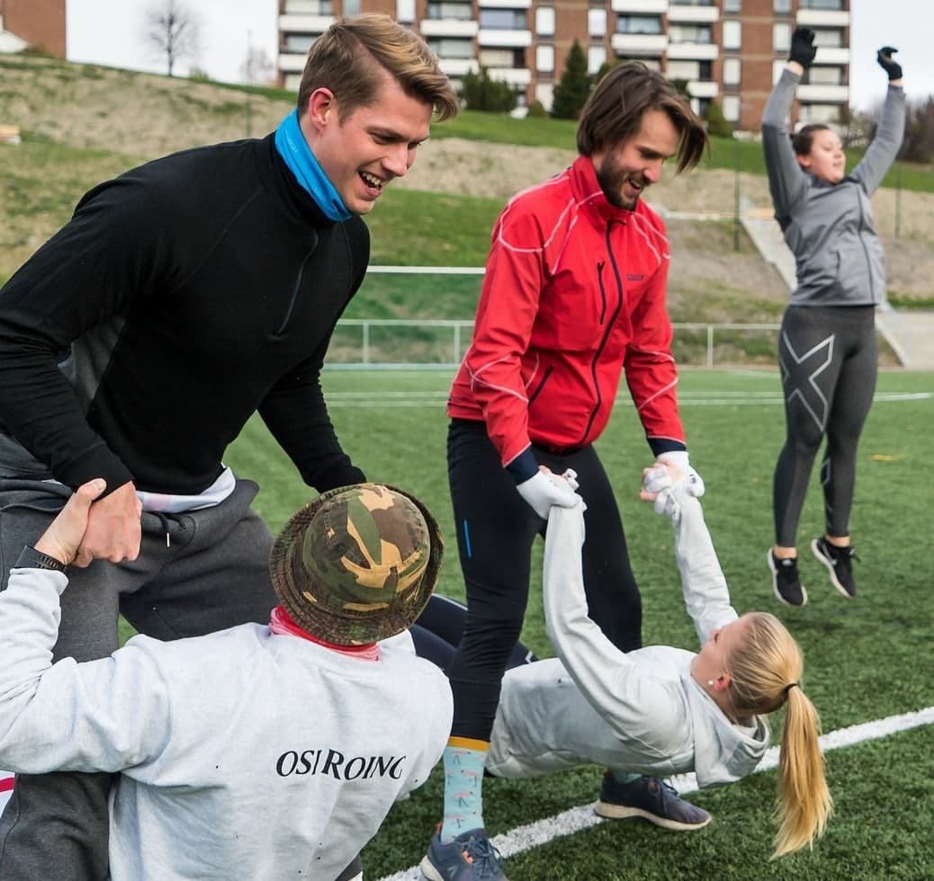Studenter trener utendørs på kunstgress; to gjør styrkeøvelser med partner mens andre smiler.