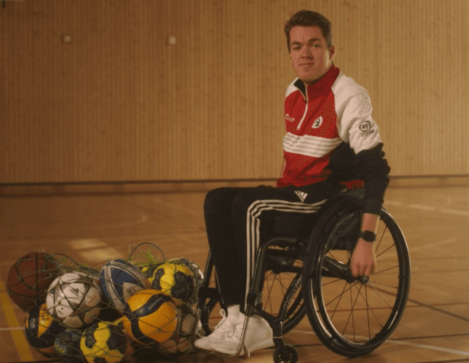 Portrett av en ung mann i rød og hvit treningsjakke som sitter i rullestol i en gymsal ved siden av et nett fulle av baller.