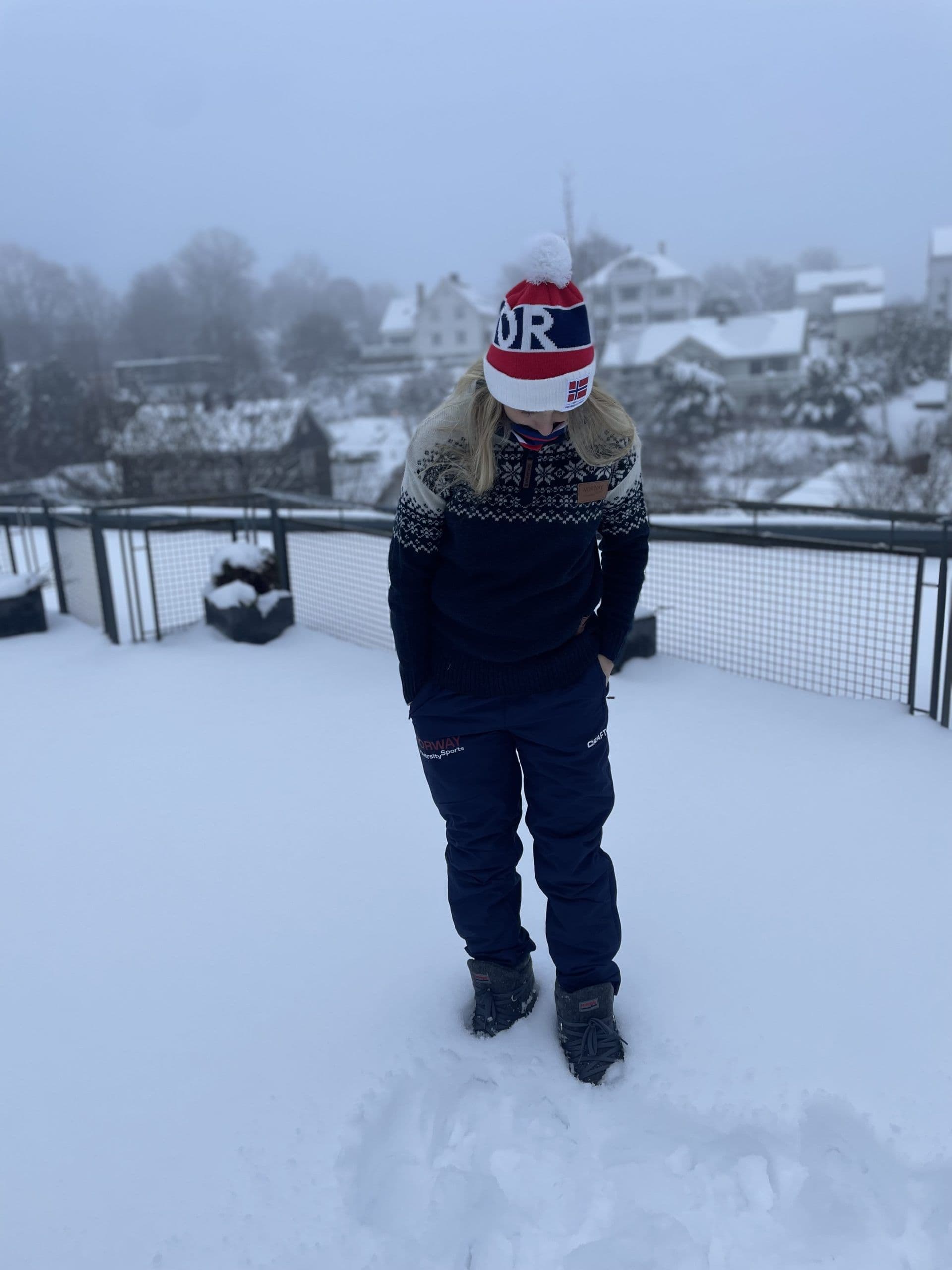 En person i vinterklær og NOR-lue står i snøen med snødekte hus i bakgrunnen.