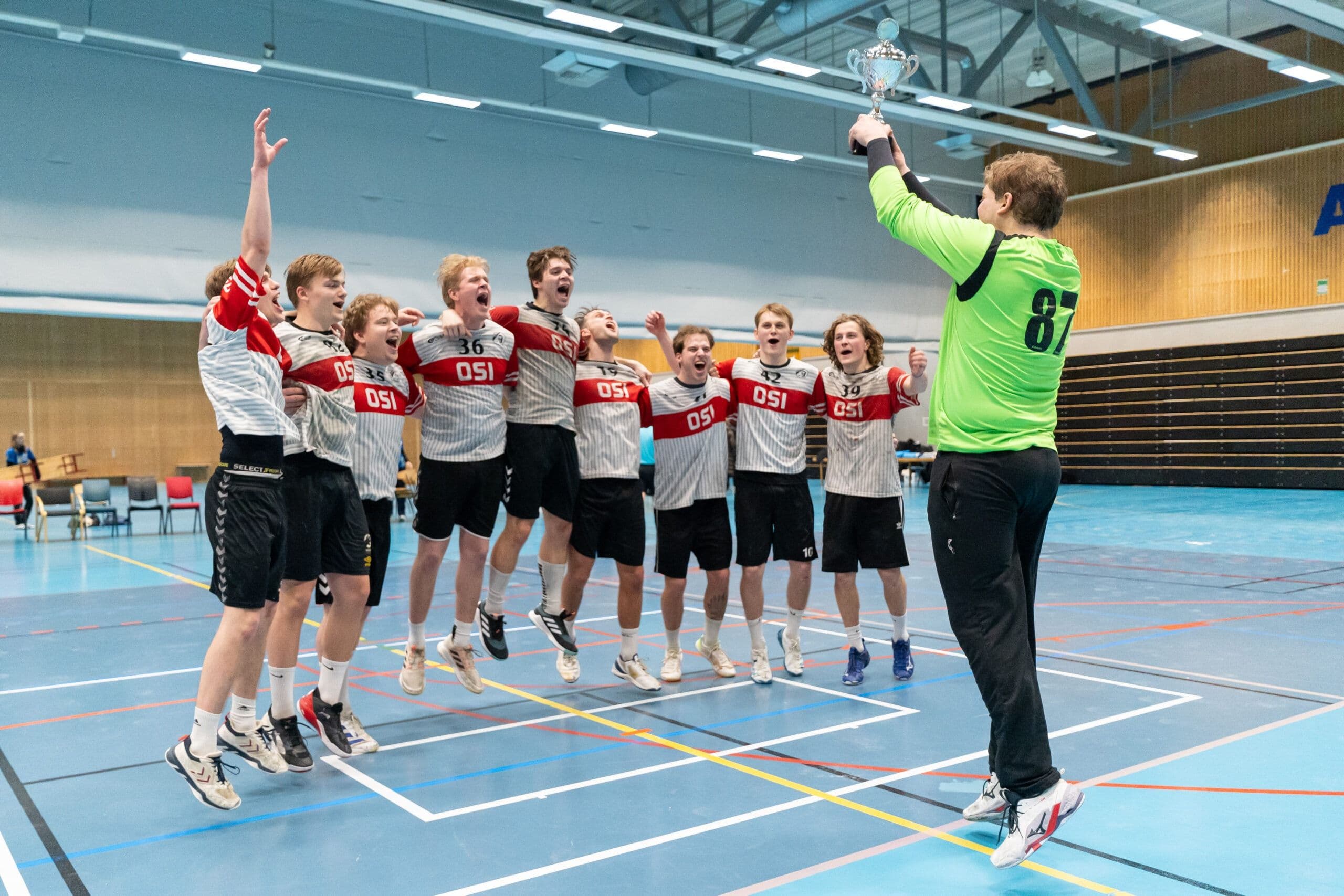 Et jublende håndballag i OSI-drakter hopper mens en spiller løfter en pokal i idrettshallen.