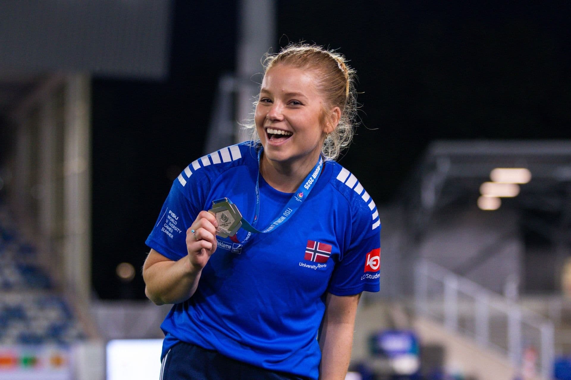 En smilende kvinne i blå drakt holder opp en medalje med norsk flagg på ermet.