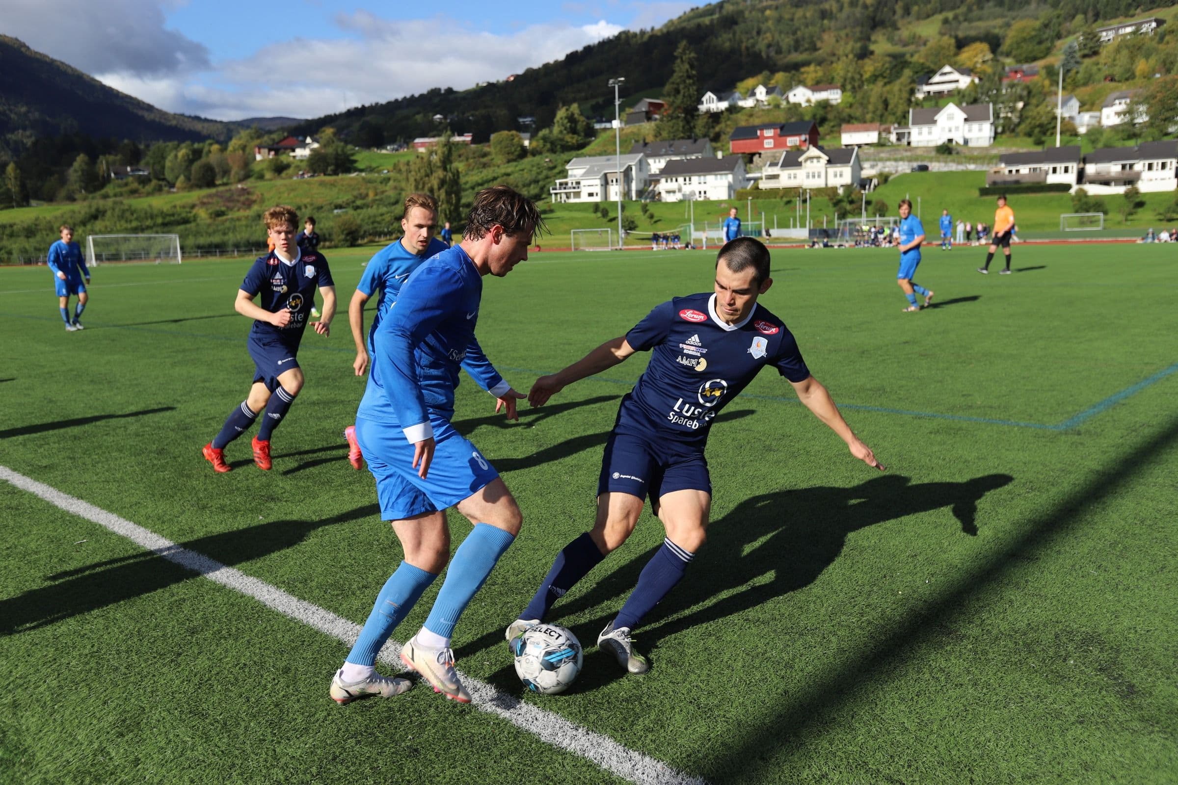 To fotballspillere i blått og mørkeblått kjemper om ballen på en kunstgressbane med fjell og hus i bakgrunnen.