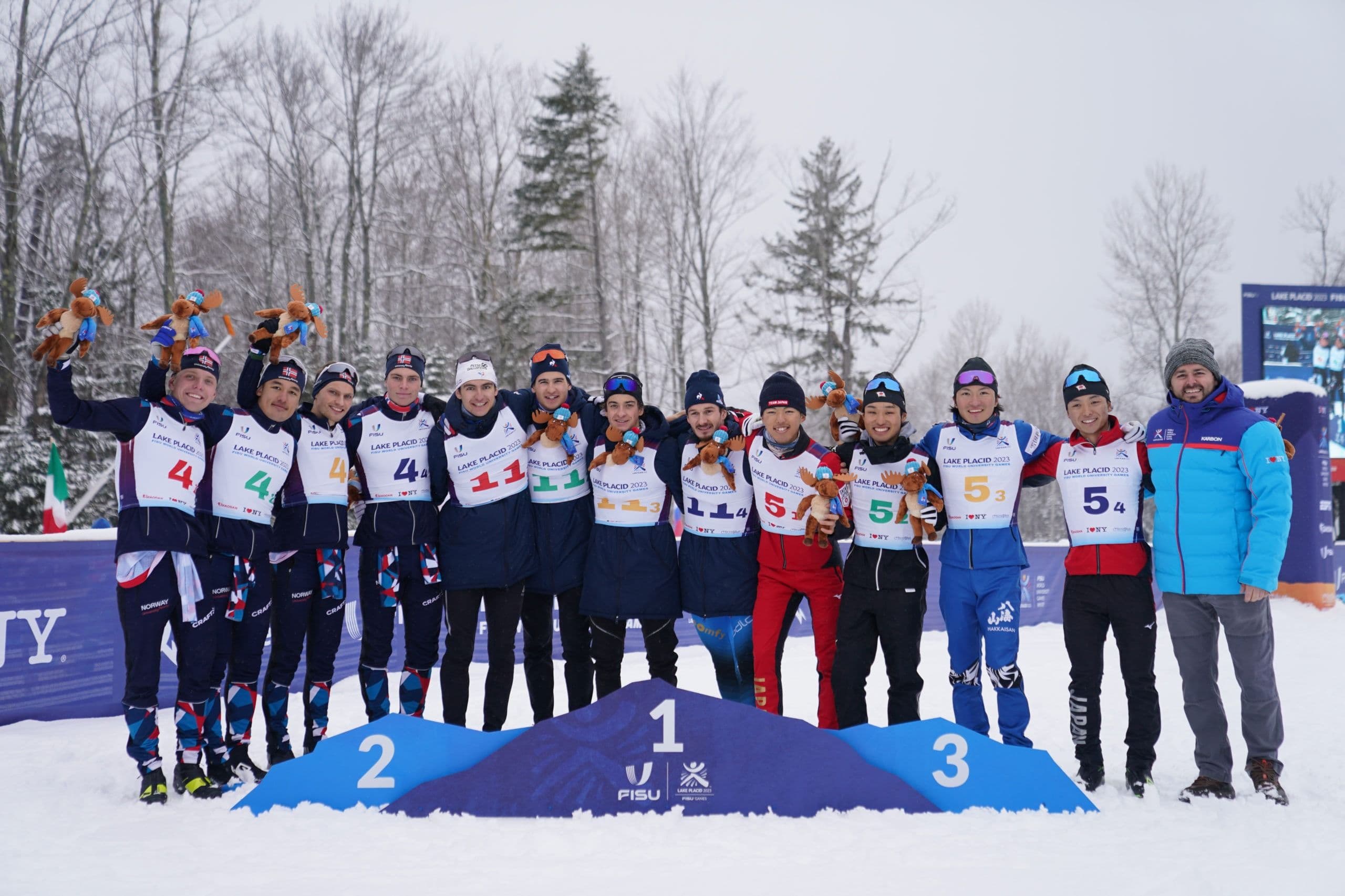 Et langrennslag jubler med troféer på premiepallen i et snødekt landskap under et universitetsmesterskap.