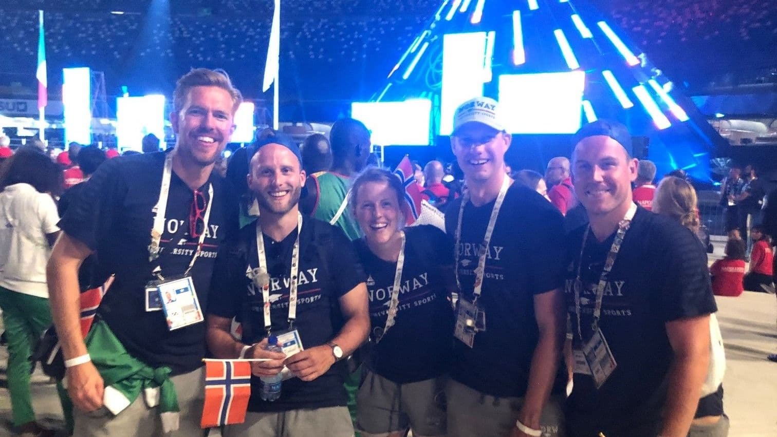 Fem personer i Norway-t-skjorter poserer sammen på en opplyst arena under en åpningsseremoni.