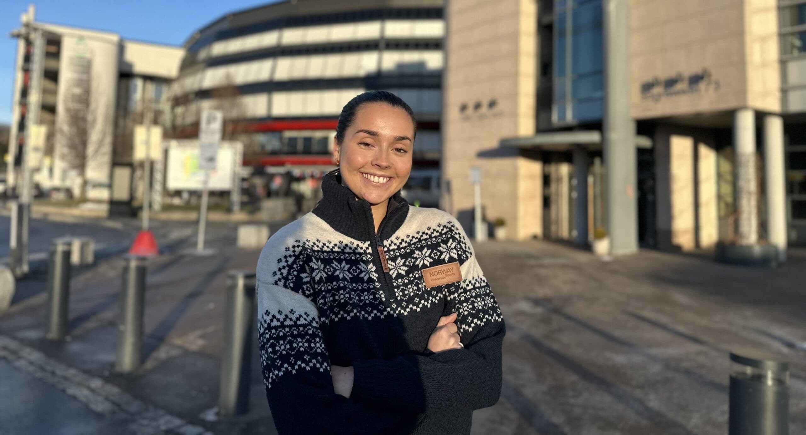 Portrett av en smilende kvinne i mønstret ullgenser utendørs foran et bysentrum i sollys.