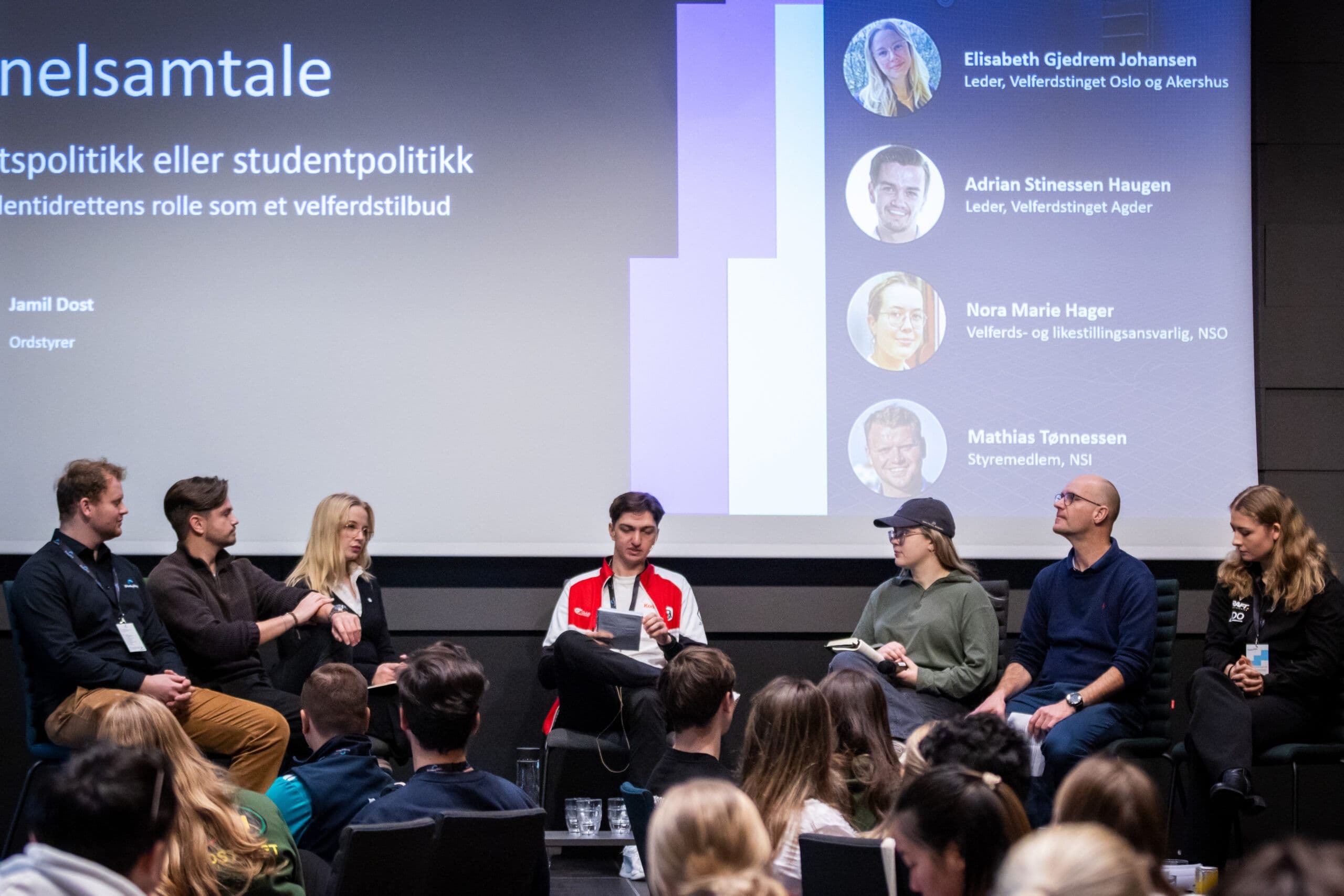 En paneldebatt på scenen der flere personer sitter i sofagruppe foran et publikum, med portretter av panelet på storskjerm.