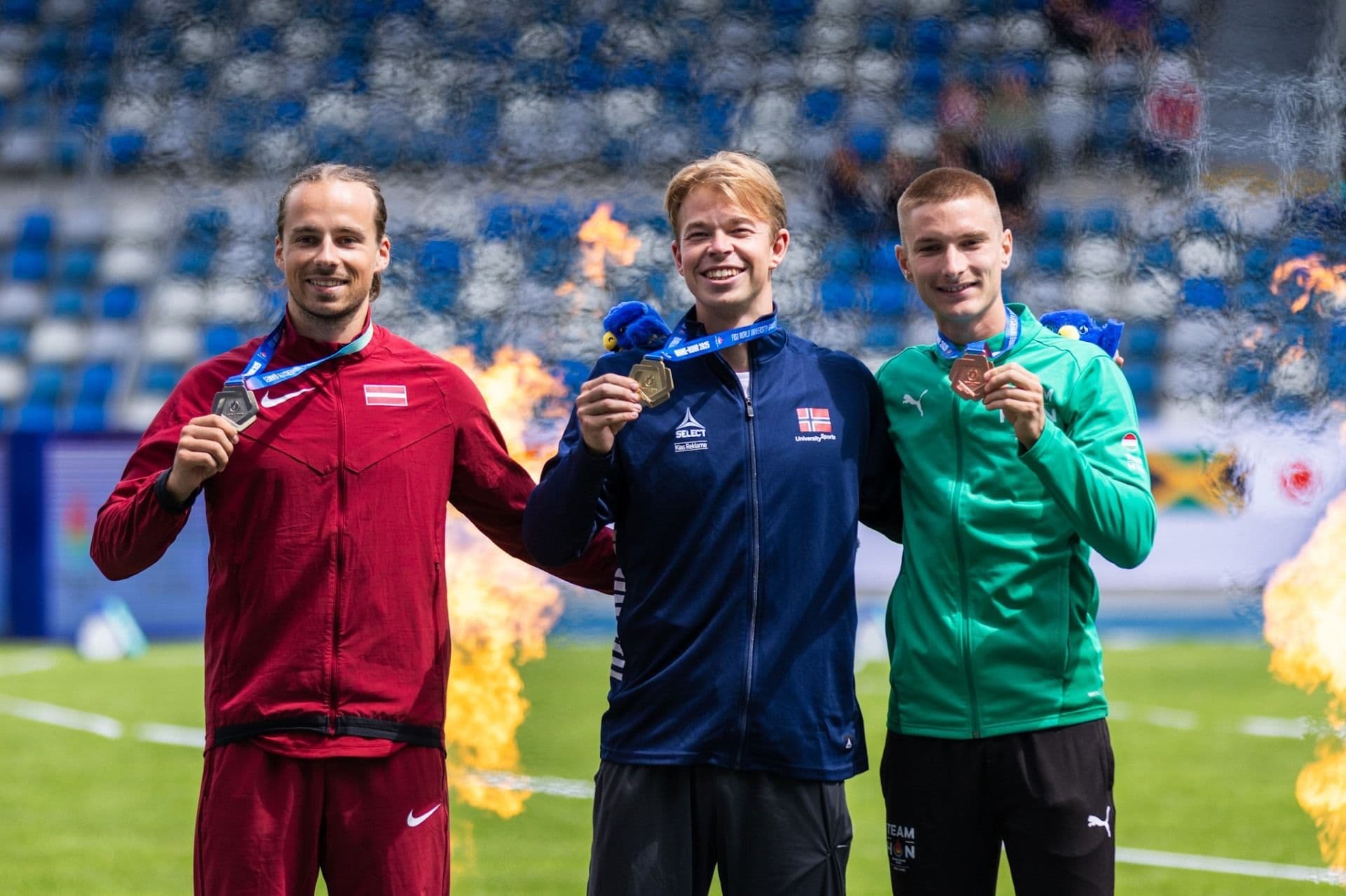 Tre mannlige friidrettsutøvere med medaljer poserer på premiepallen med flammer i bakgrunnen.