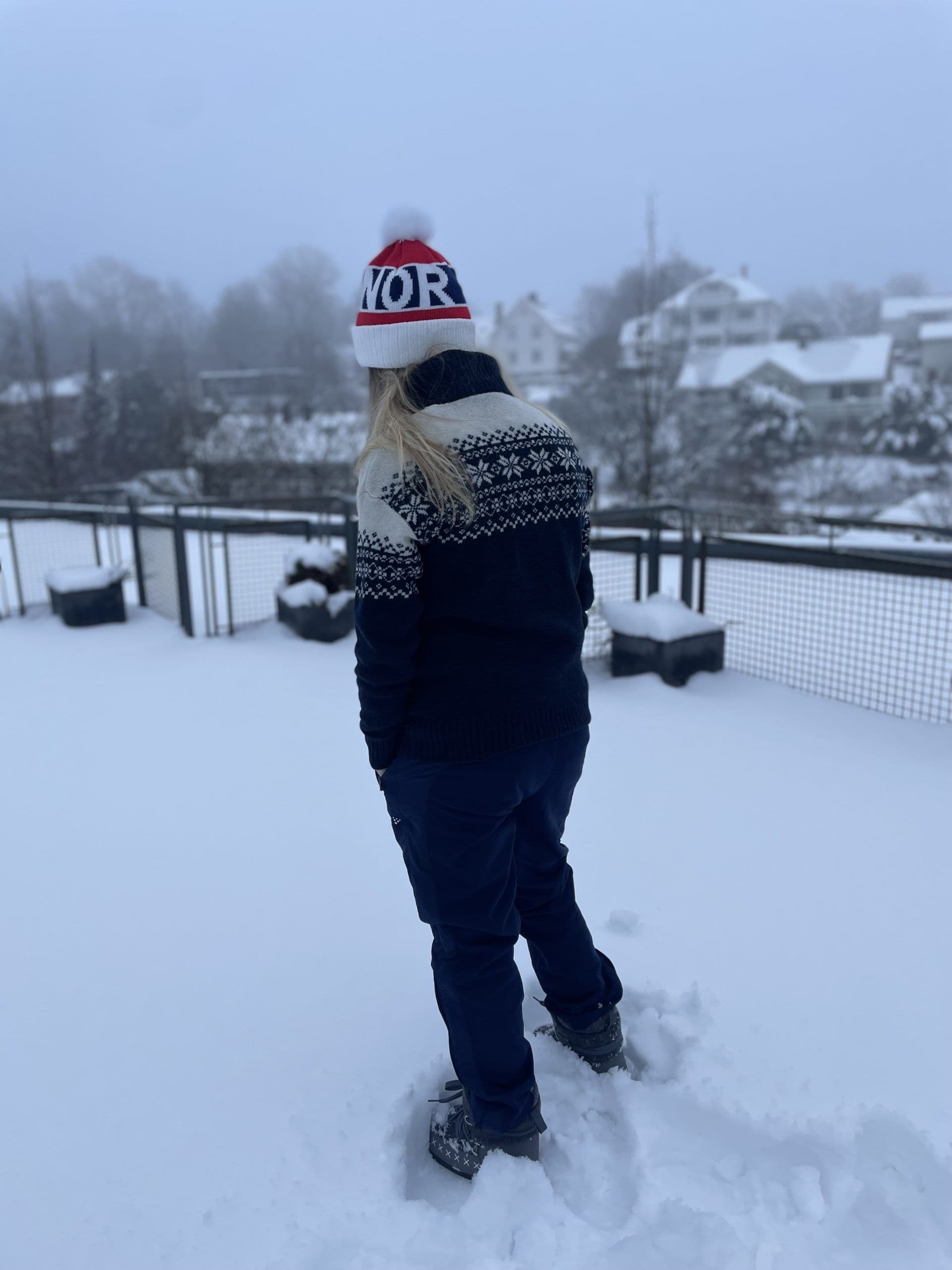 En person i mønstret genser og NOR-lue står med ryggen til i snøen med snødekte hus i bakgrunnen.