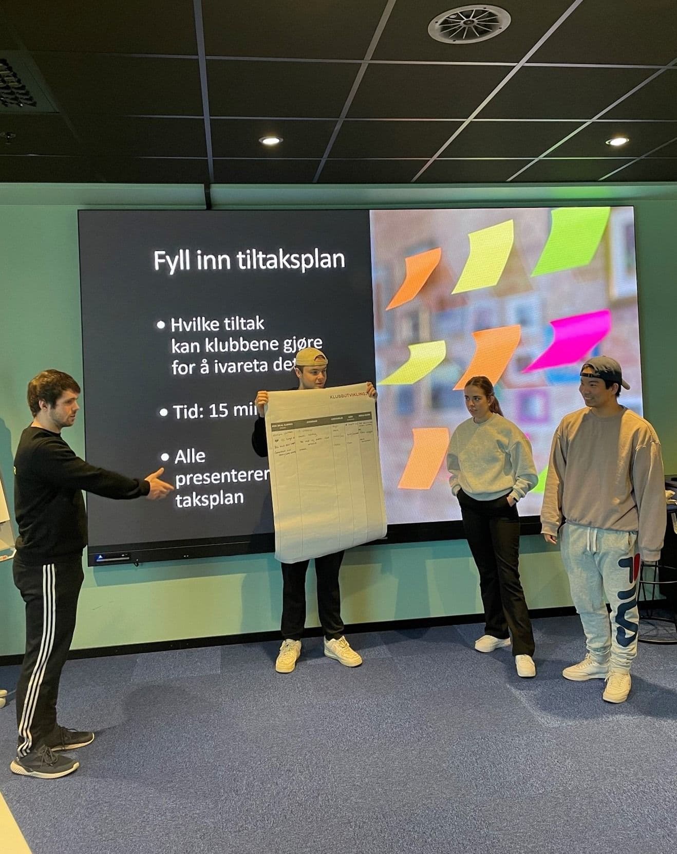 Studenter presenterer foran en skjerm med teksten «Fyll inn tiltaksplan» under en workshop.