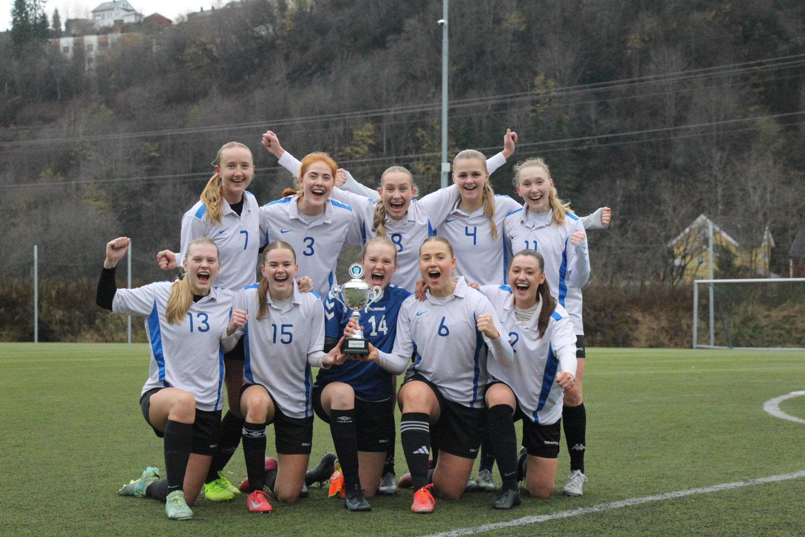 Et kvinnelig fotballag jubler med pokal på kunstgressbanen.