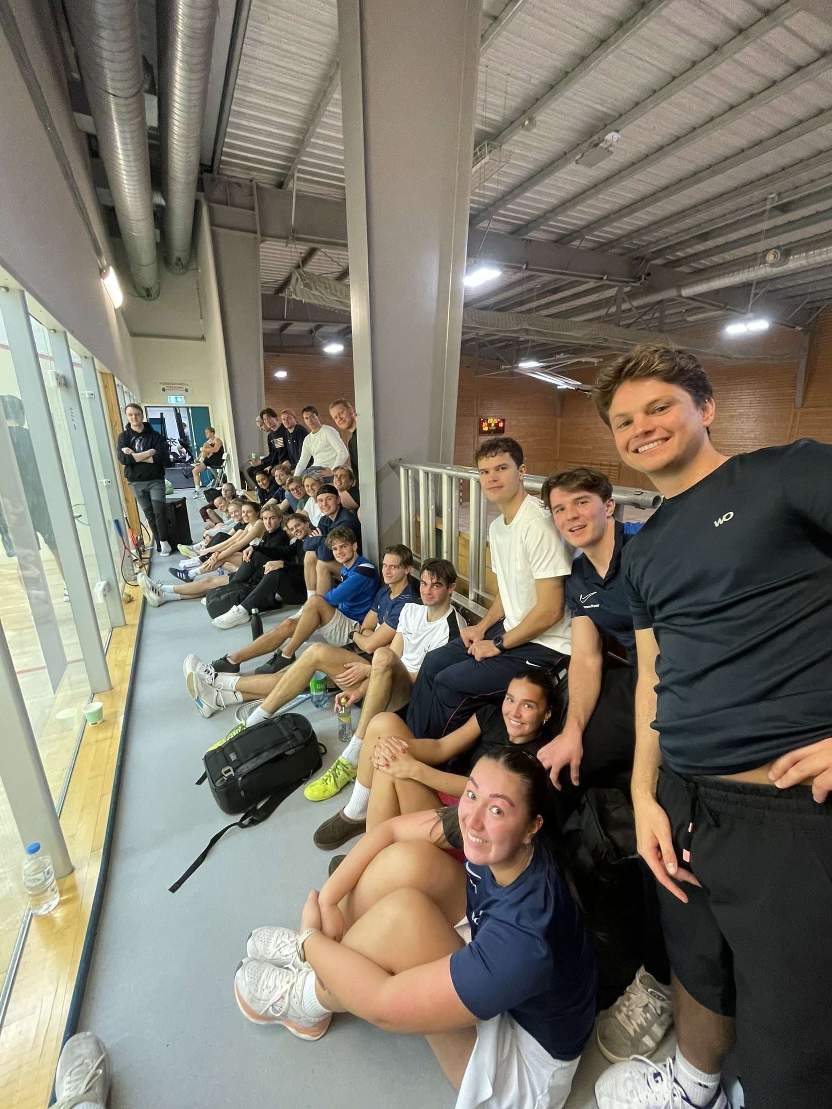 En gruppe studenter sitter langs galleriet ved en squashbane.