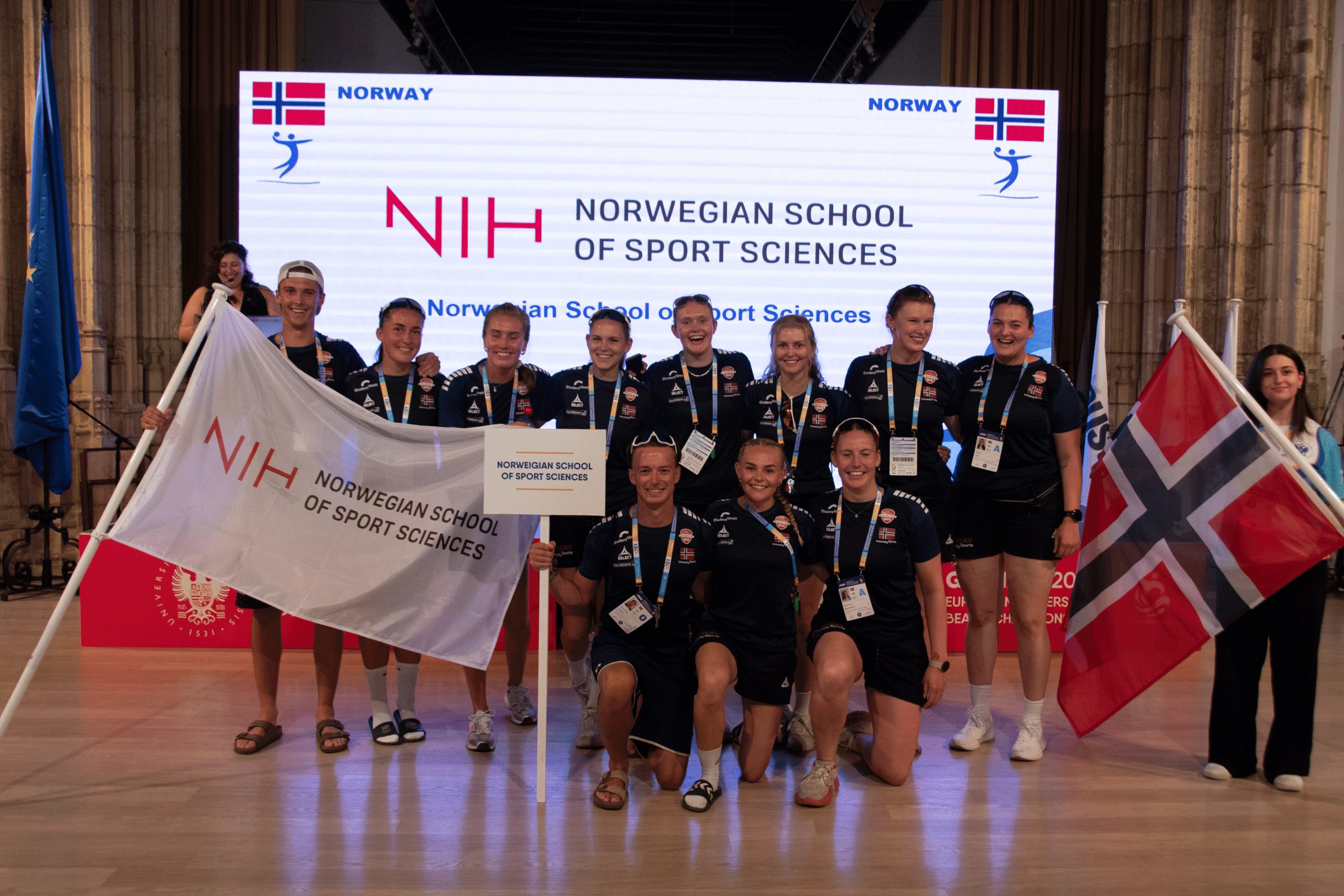 En gruppe utøvere poserer med flagg og bannere for Norges idrettshøgskole (NIH) under et arrangement.