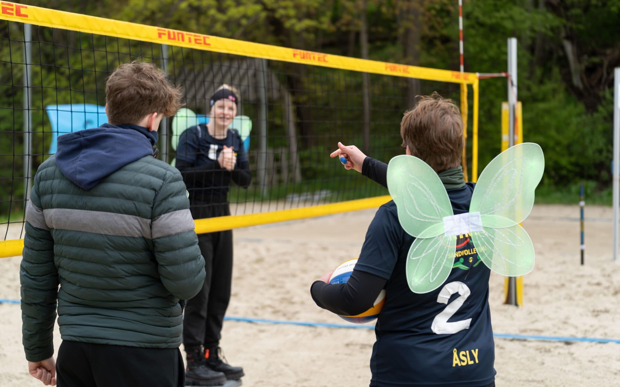 Ungdommer spiller sandvolleyball, der en av spillerne har på seg grønne feevinger.