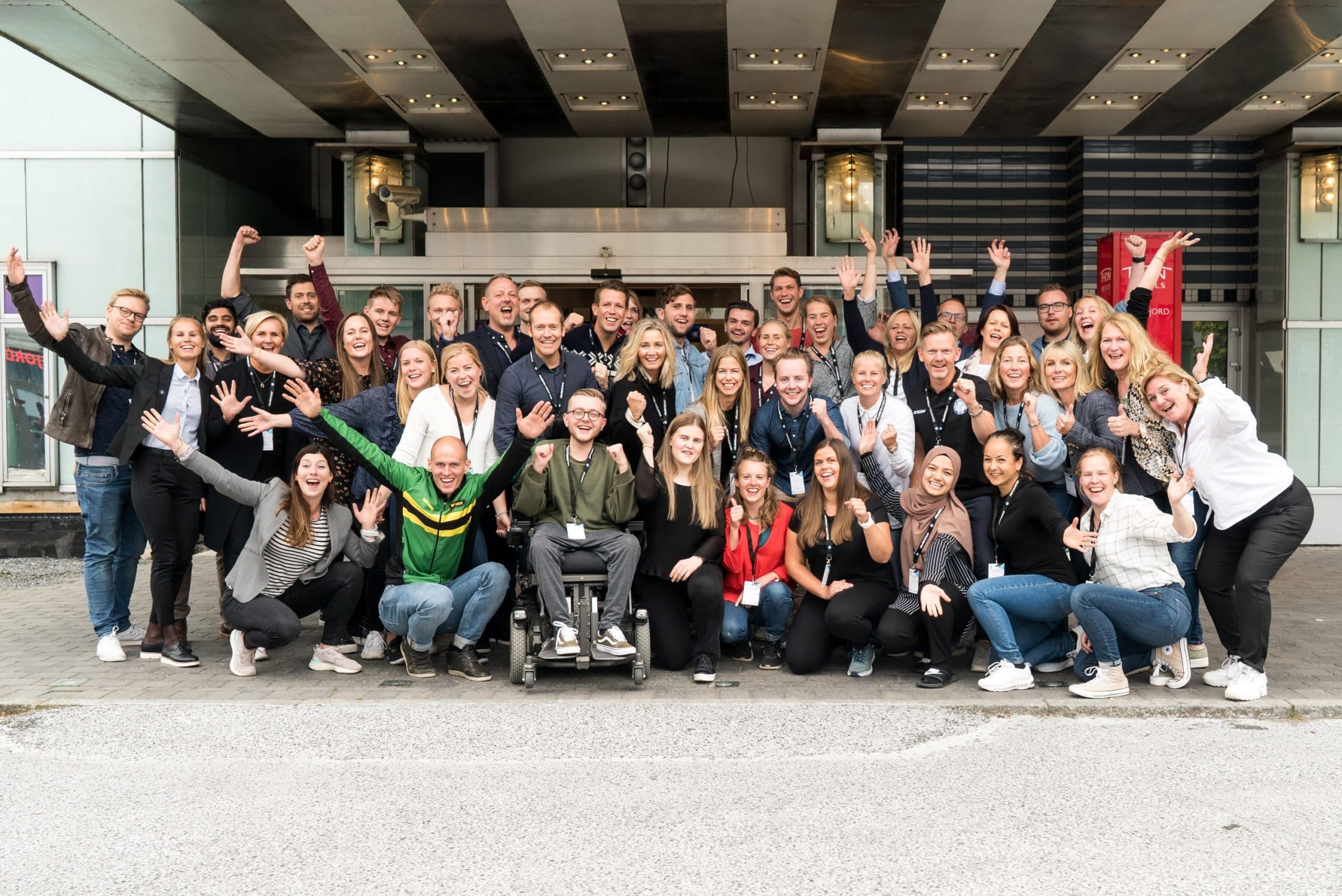 En stor gruppe mennesker jubler foran en bygning, med en person i rullestol midt i bildet.