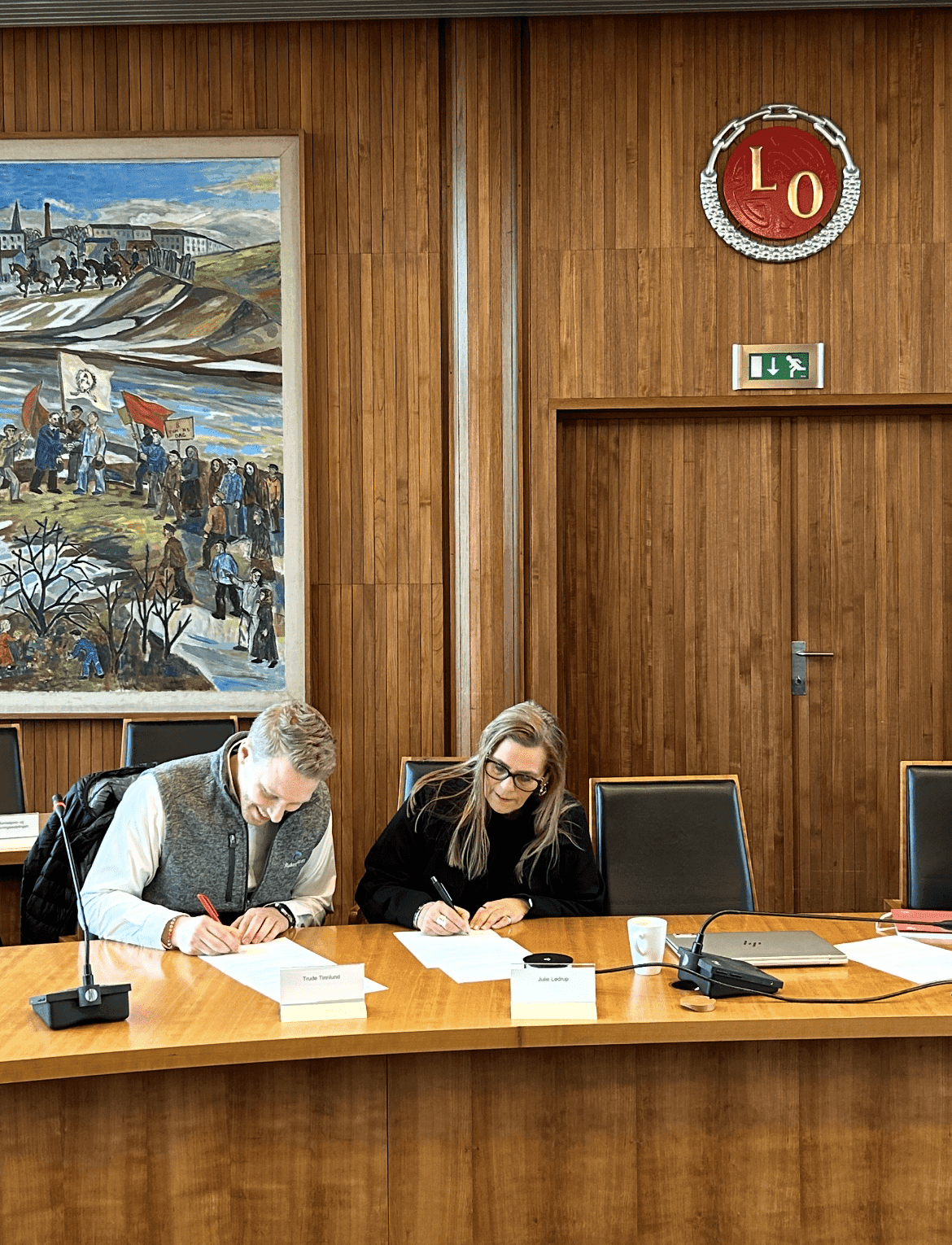 En mann og en kvinne signerer dokumenter ved et møtebord i et rom med LO-logo på veggen.