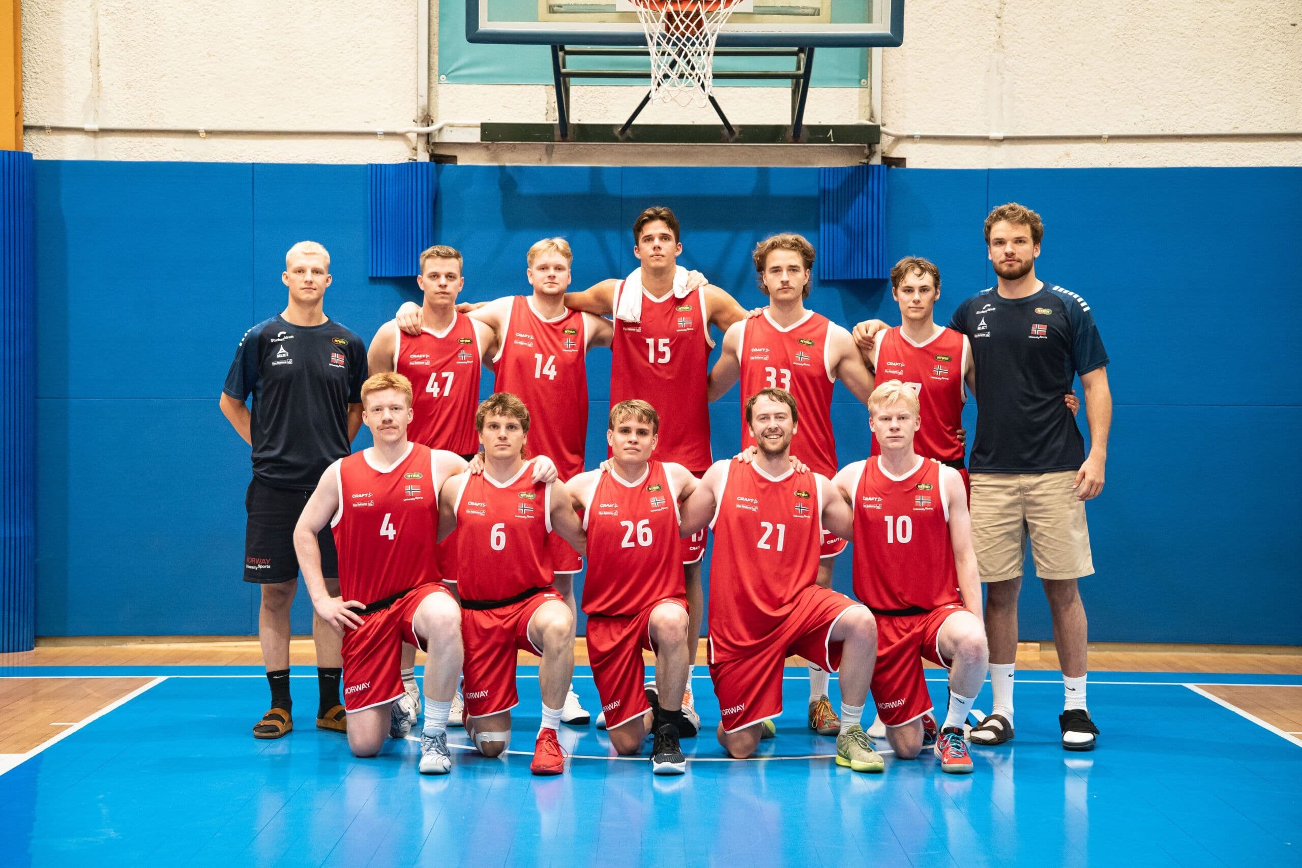 Et lag med mannlige basketballspillere i røde drakter poserer sammen med to trenere i en idrettshall.