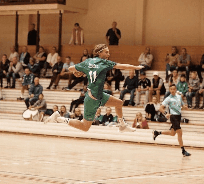 En håndballspiller i grønn drakt med nummer 11 svever i lufta på vei mot mål foran tribunen.