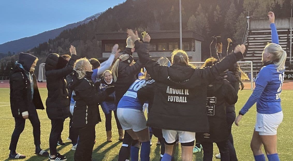 Et fotballag jubler i en klynge på en fotballbane i skumringen, en spiller med teksten Årvoll IL Fotball på ryggen.
