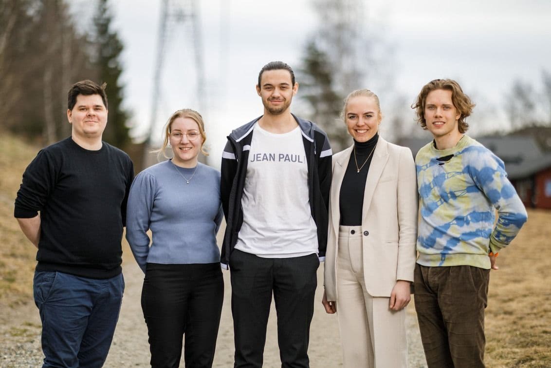 Fem unge voksne står oppstilt side om side på en grusvei i et landlig landskap.