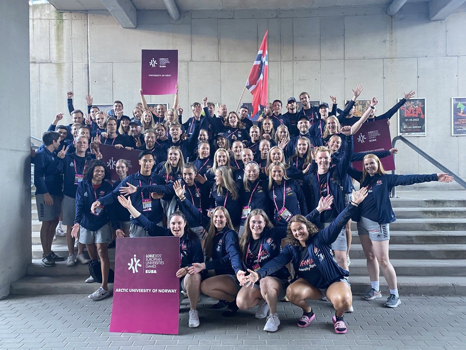 En stor gruppe studenter i blå overdeler jubler foran et norsk flagg på en trapp under et europeisk studentmesterskap.