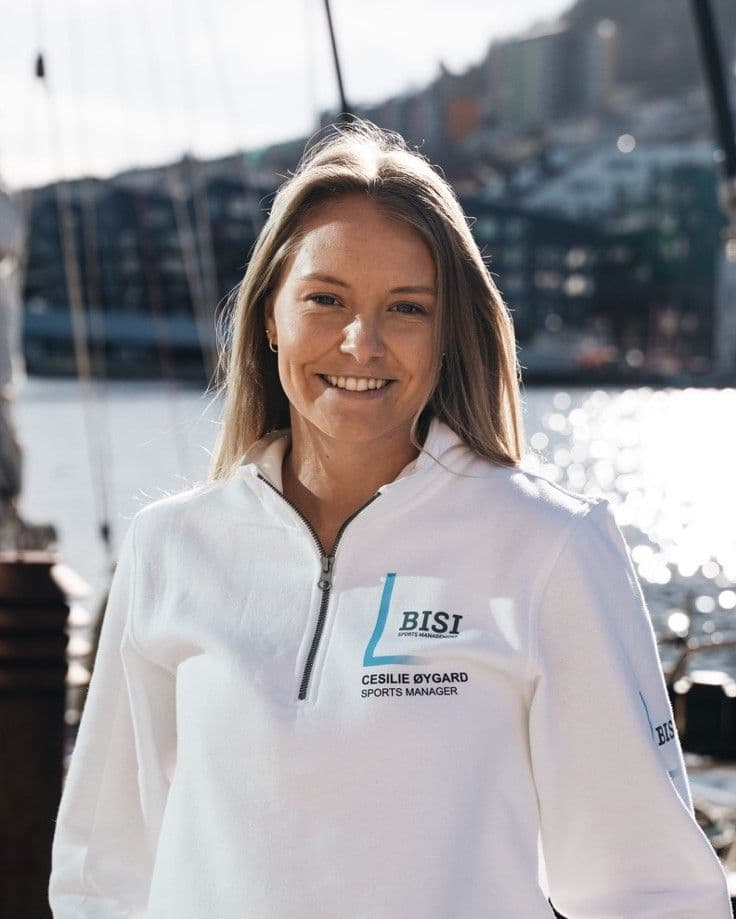 Portrett av en smilende kvinne i hvit genser med BISI-logo og teksten Sports Manager, foran en havn.