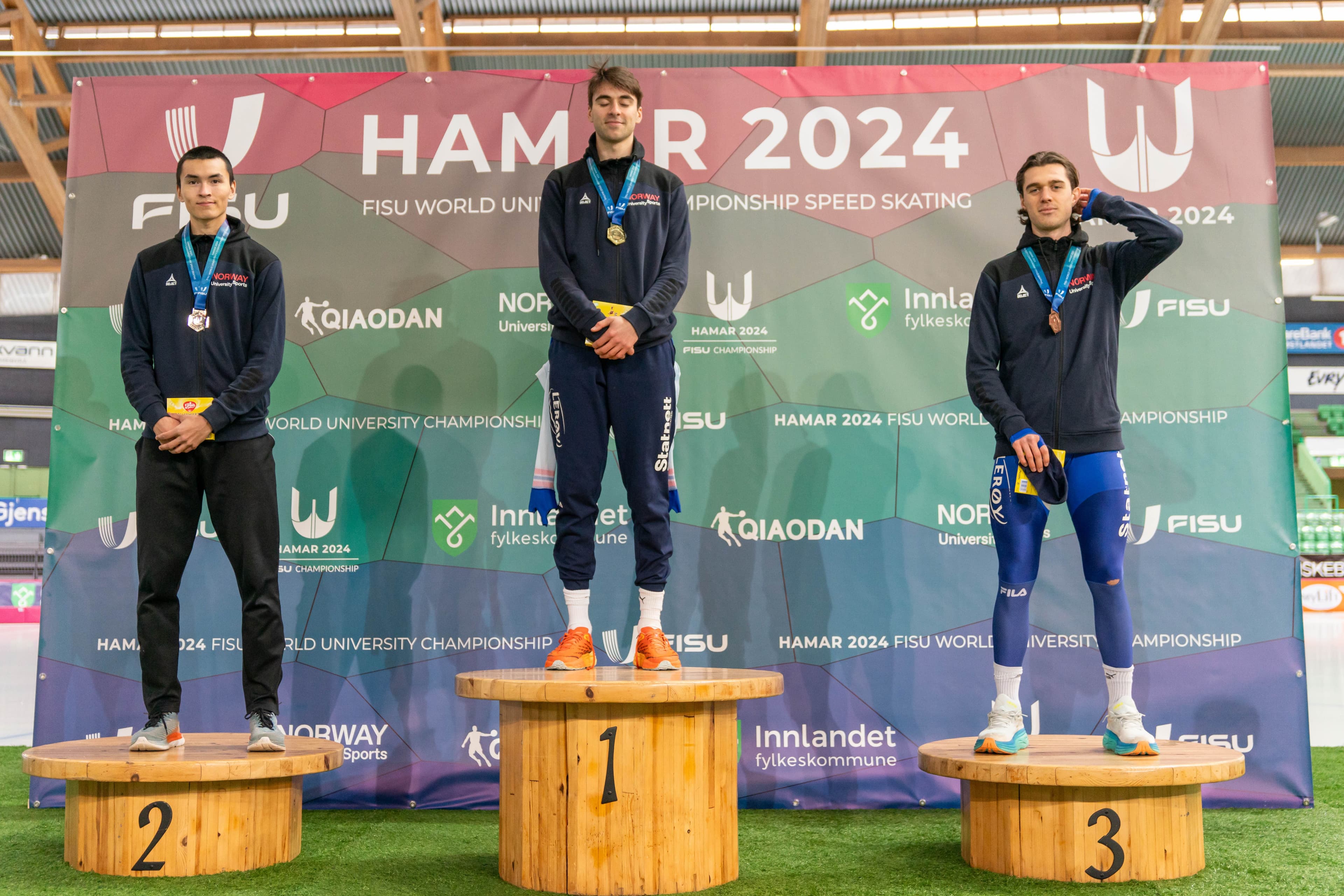 Tre skøyteløpere med medaljer på premiepallen foran veggen for Hamar 2024 FISU World University Championship Speed Skating.