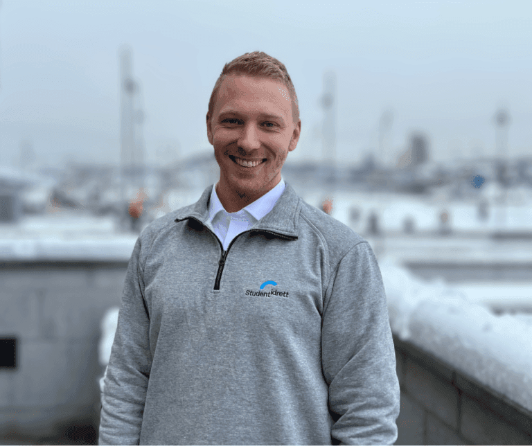 Portrett av en smilende mann i grå Studentidrett-genser på en snødekt takterrasse.
