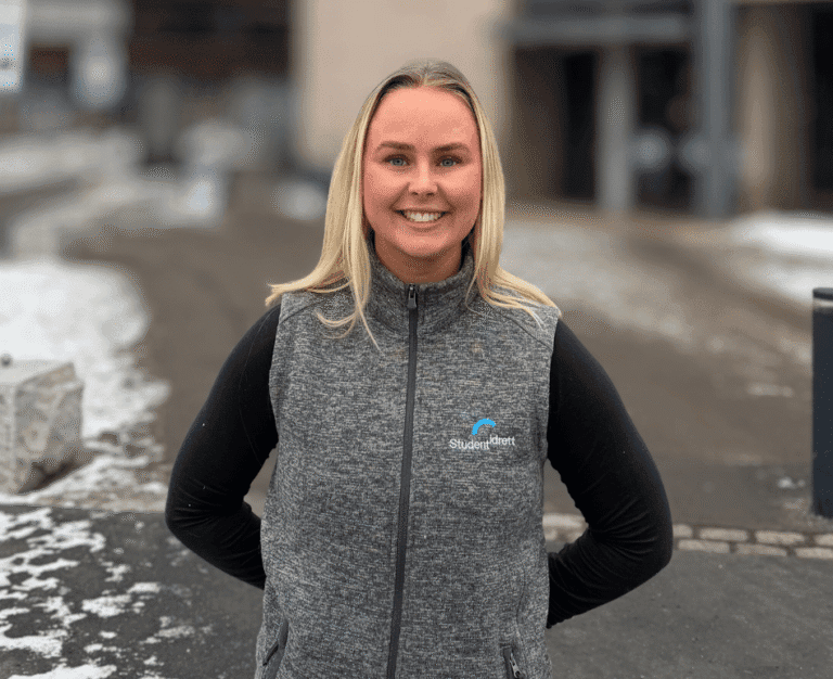 Portrett av en smilende kvinne i grå Studentidrett-vest på et snødekt uteområde.