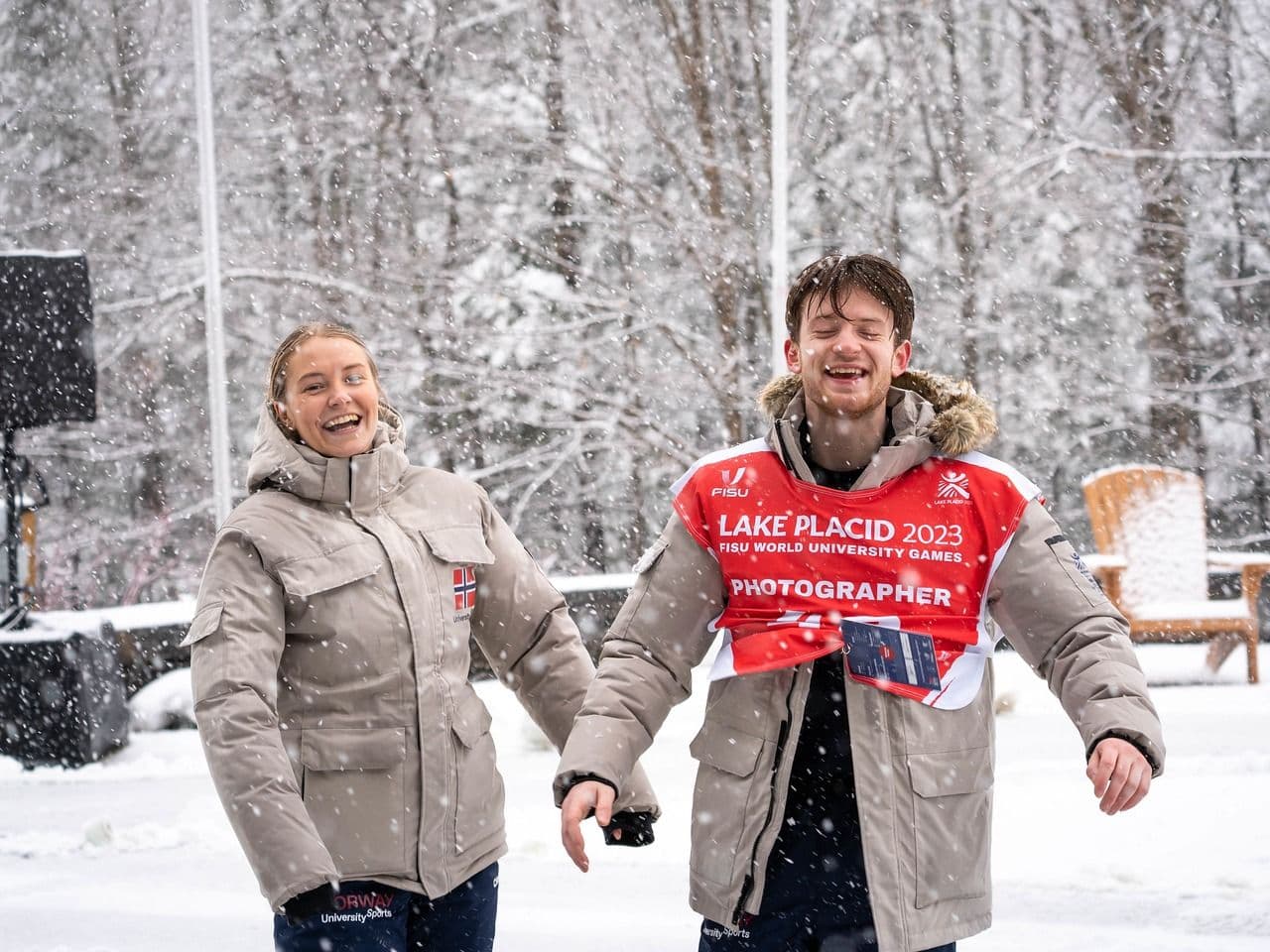 En kvinne og en mann ler i snøvær, mannen i jakke med teksten Lake Placid 2023 Photographer.