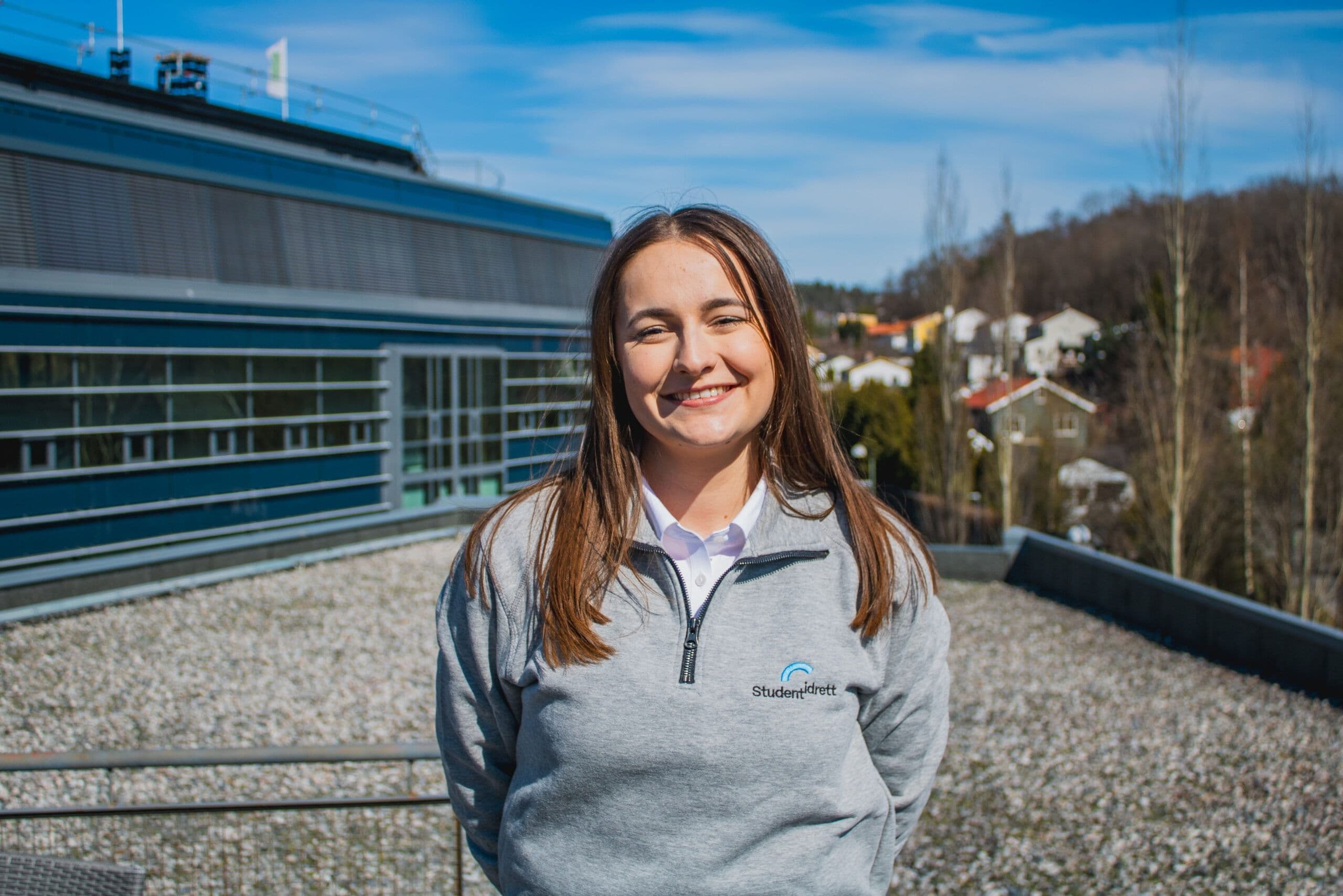Portrett av en kvinne i grå Studentidrett-genser på et tak med utsikt over et boligområde.