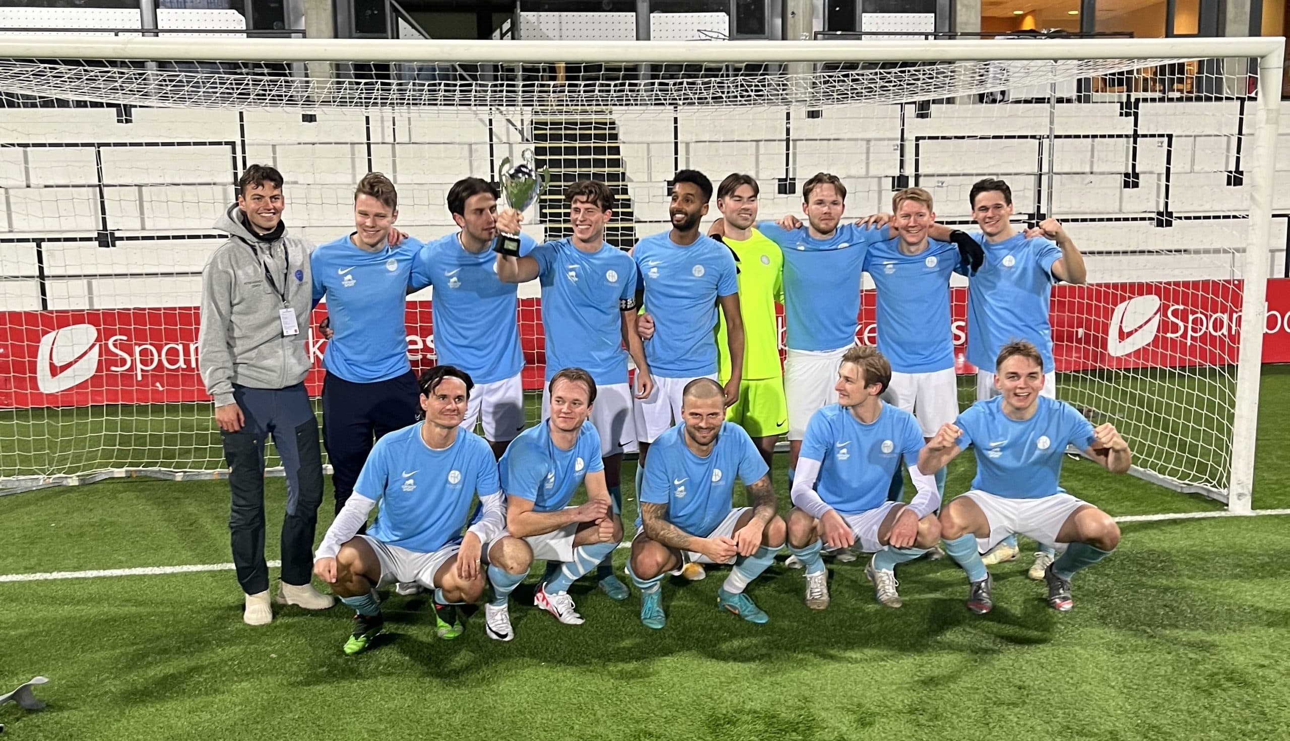 Et fotballag i lyseblå drakter poserer med en pokal foran et mål.