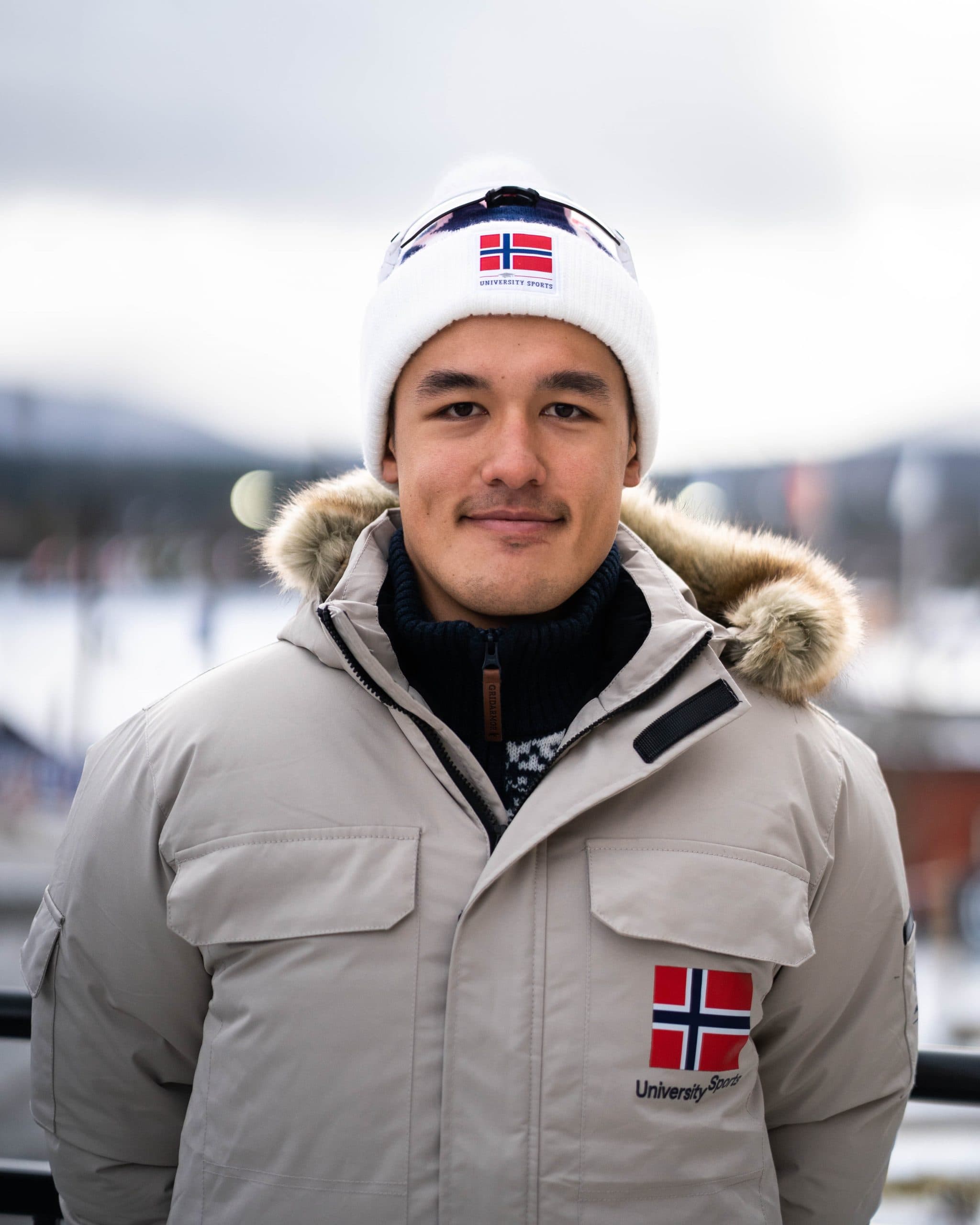 Portrett av en mann med hvit lue og jakke med norsk flagg, foran et vinterlandskap.