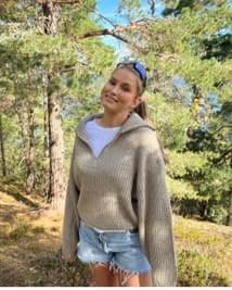 En kvinne i beige genser står i et skogsområde en solrik dag.