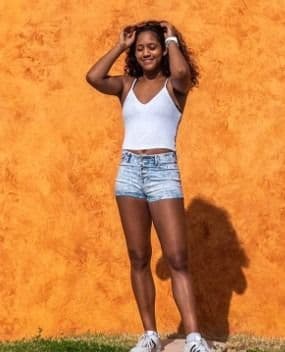 En kvinne står foran en oransje vegg iført hvit topp og shorts.