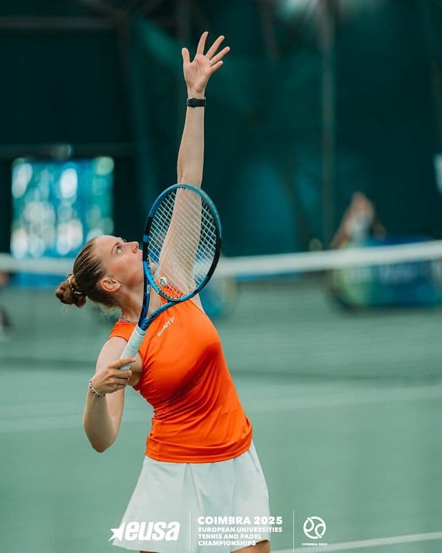 En tennisspiller i oransje overdel utfører en serve på en innendørs bane.