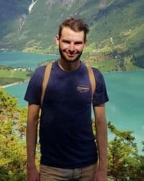 Portrett av en mann i mørk t-skjorte foran en fjord og fjellandskap.