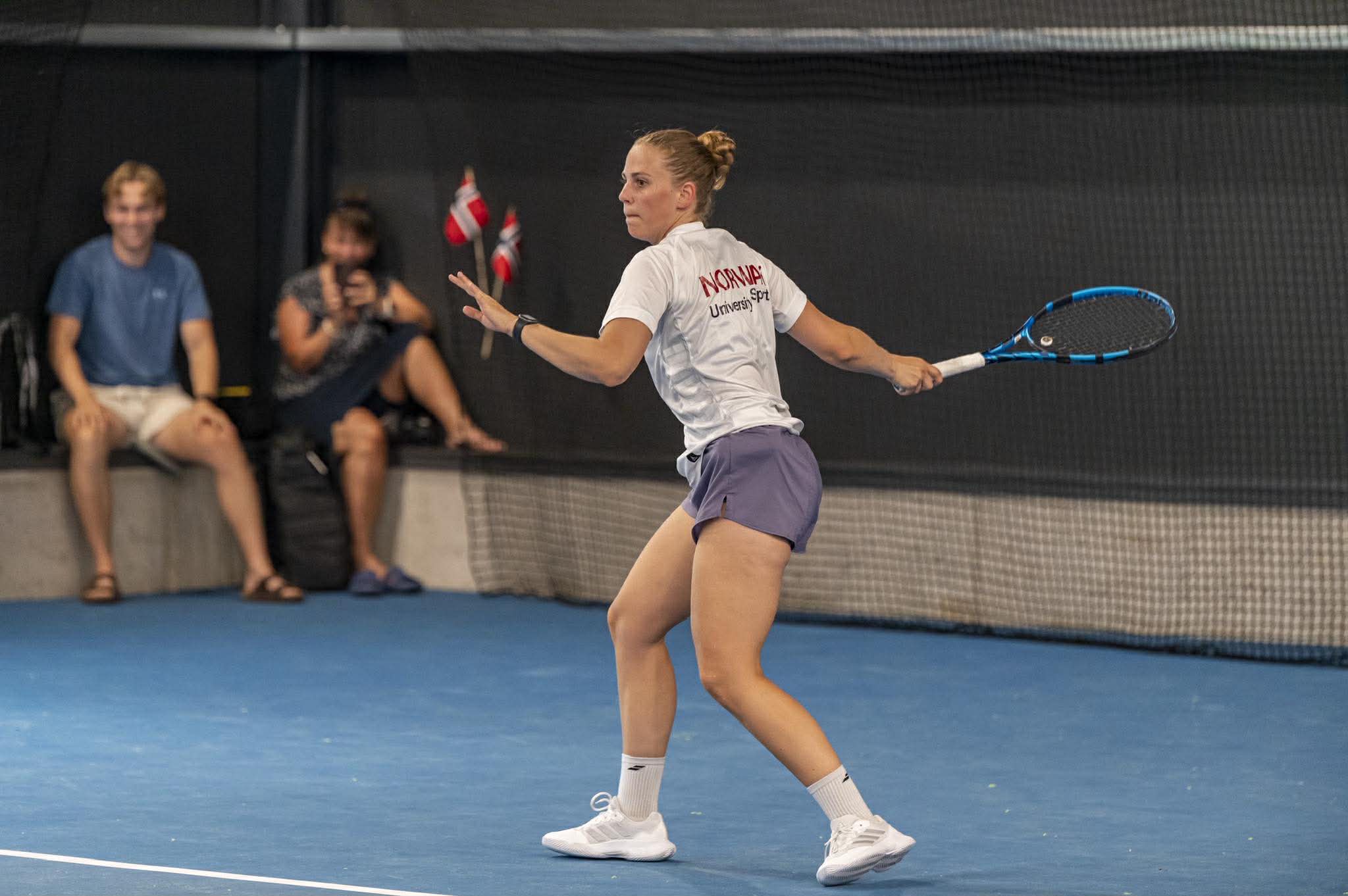 En tennisspiller i hvit trøye slår en forhånd på en innendørs bane.