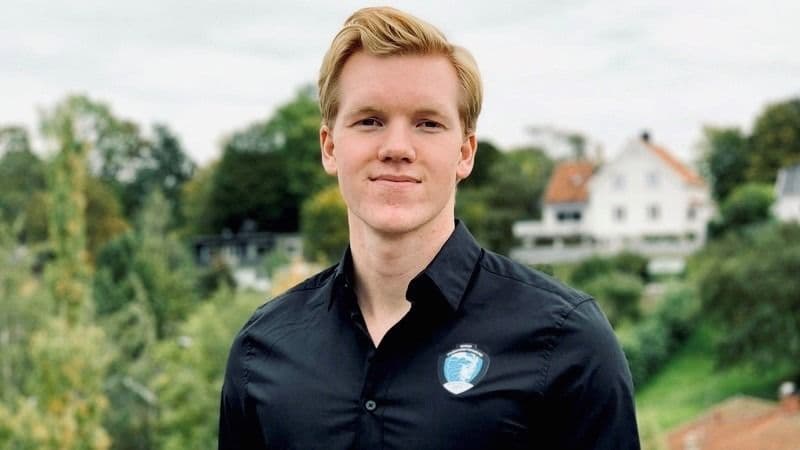 Portrett av en ung mann i mørk skjorte utendørs med grønt landskap i bakgrunnen.