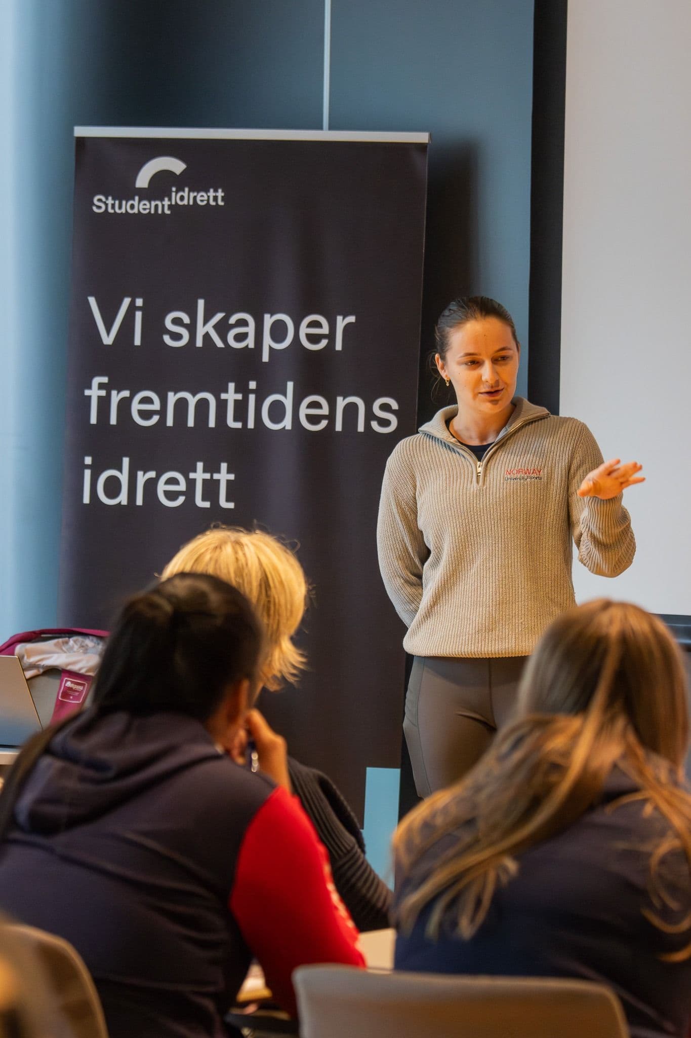 En kvinne holder et foredrag foran en Studentidrett-rollup med teksten «Vi skaper fremtidens idrett».