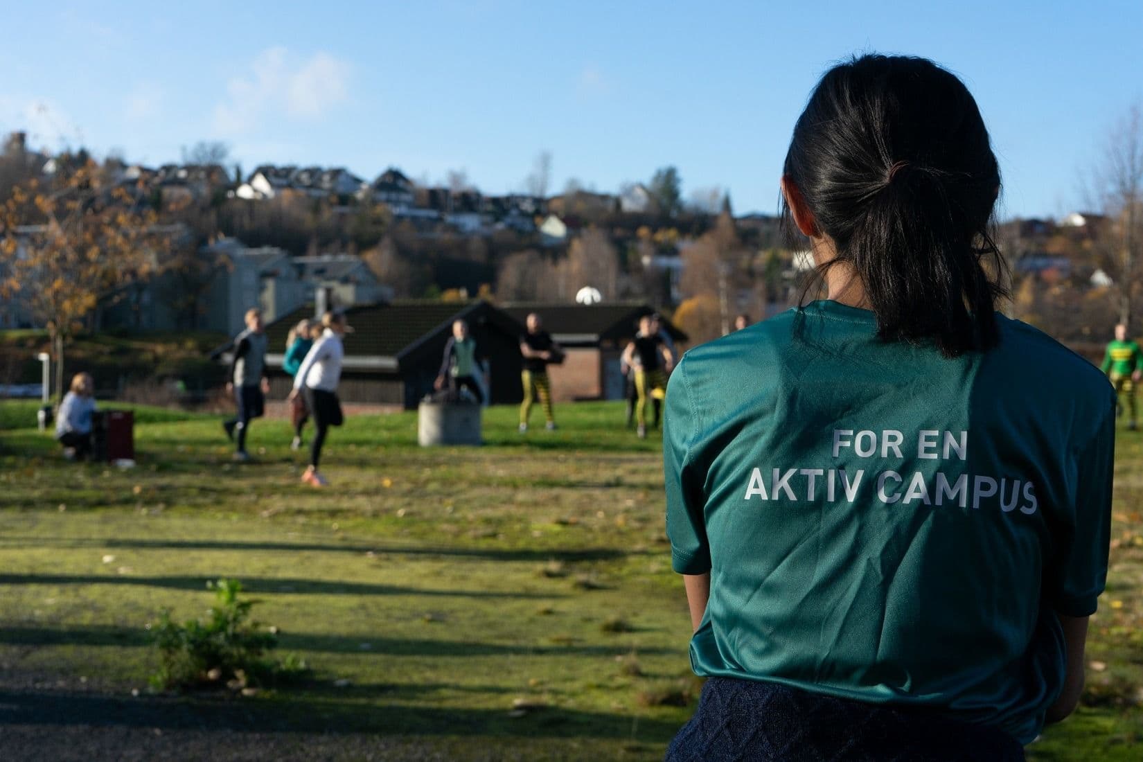 En person i grønn t-skjorte med teksten «For en aktiv campus» ser på folk som trener i en park.
