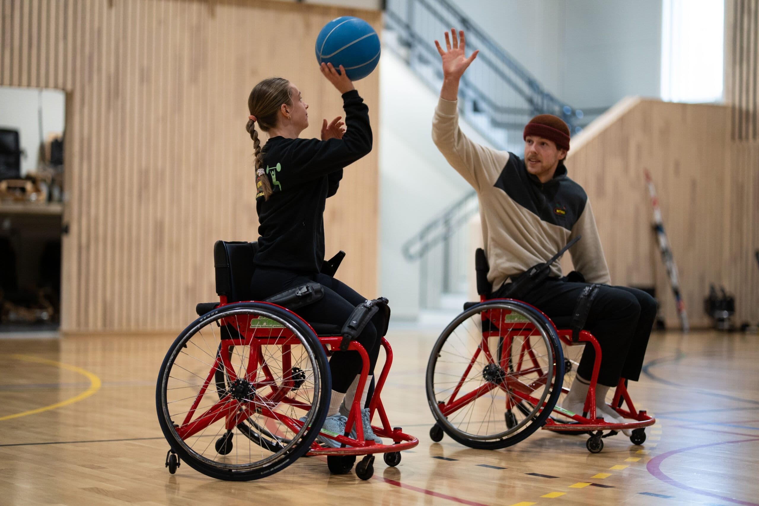 To personer i rullestol spiller basketball i en idrettshall.