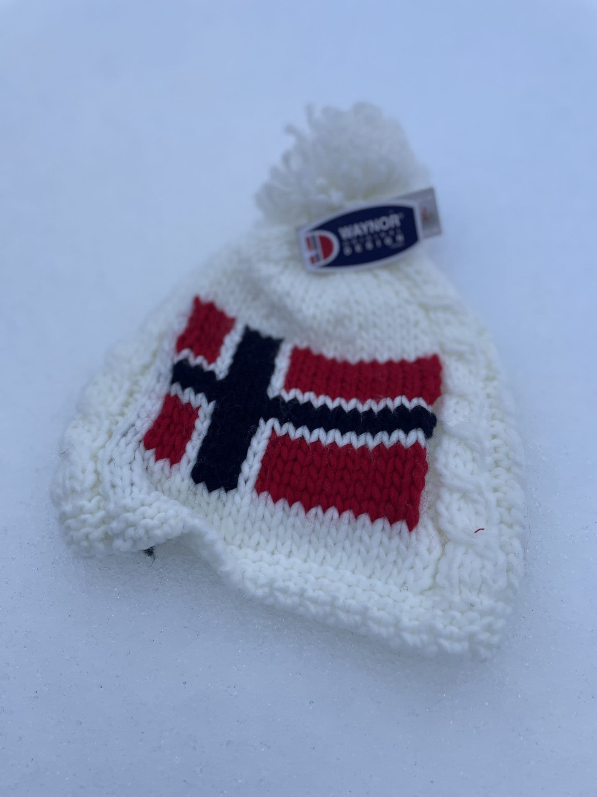 En hvit strikkelue med norsk flagg og dusk ligger på snøen.