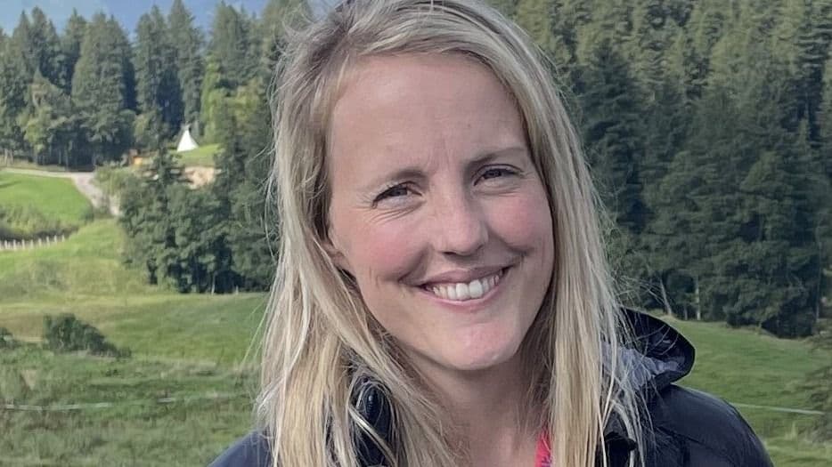 Portrett av en smilende blond kvinne utendørs med grønt kupert landskap i bakgrunnen.