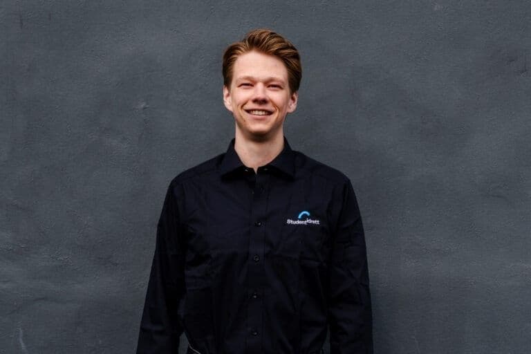 Portrett av en smilende mann i mørk skjorte med Studentidrett-logo foran en grå vegg.