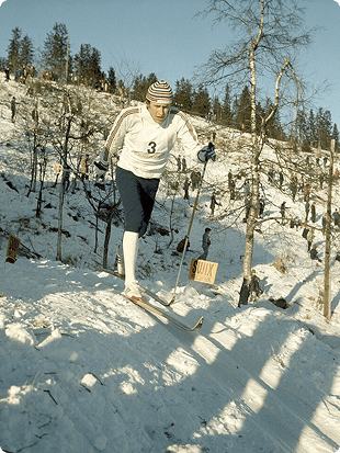 Eldre fargefoto av en langrennsløper med startnummer 3 som går motbakke i et snødekt terreng.