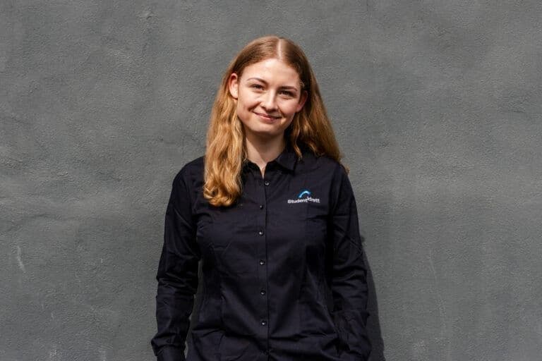Portrett av en smilende kvinne i mørk skjorte med Studentidrett-logo foran en grå vegg.